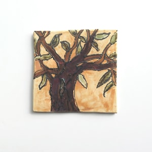 May include: Square ceramic tile featuring a stylized tree design. The tree has a dark brown trunk and branches with green leaves. The background is a light tan color, and the tile has a slightly textured surface.