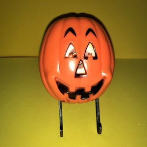 May include: A bright orange plastic jack-o'-lantern decoration with a carved face featuring triangular eyes and a wide, toothy grin outlined in black. The pumpkin is supported by two thin, black metal legs that extend downward from its base.