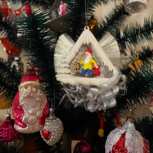 May include: Christmas tree ornaments, including a Santa Claus figure and a whimsical gnome carrying a lantern. The gnome is inside a snowy, triangular structure with a white fringe. Other ornaments are visible in the background.