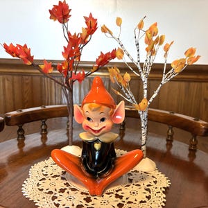 May include: A ceramic elf figurine with a pointed orange hat, large ears, and a black outfit featuring a gold heart emblem. The elf sits in a cross-legged position between two miniature decorative trees with orange and yellow autumn leaves.