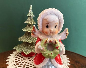 May include: A ceramic angel figurine wearing a red coat with white trim and a matching hood, holding a small green wreath with red accents. The angel has gold-tipped wings and is positioned next to a small, textured green Christmas tree.