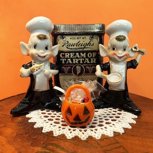May include: Two ceramic elf chef figurines wearing white hats and black uniforms with gold trim, positioned on either side of a metal Rawleigh's Cream of Tartar tin. A small orange plastic jack-o'-lantern bucket sits in the center foreground.