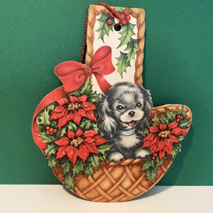 May include: A Christmas ornament featuring a black and white puppy in a woven basket filled with red poinsettias and holly. A red bow and a decorative top with holly and berries complete the design. The ornament is suspended by a red string.