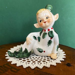 May include: A vintage ceramic elf figurine with a green cap and holly leaf and berry accents on its white outfit. The elf is seated on a white lace doily, with a green background.