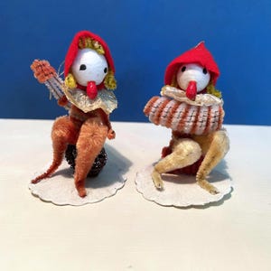 May include: Two vintage doll figures. Each doll has a white head, red hood, and a ruffled collar. One doll holds a musical instrument. The dolls are made of fabric and are on a white paper base.