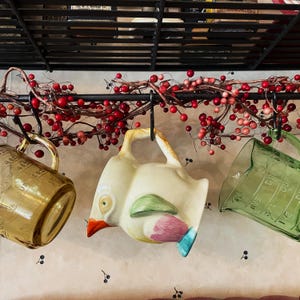 May include: Three hanging kitchen items: a translucent amber glass measuring cup with embossed markings, a cream-colored ceramic bird-shaped pitcher with a bright orange beak and painted wing details, and a translucent green glass measuring cup.