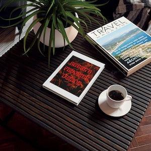 May include: A white book with the title "Satan's Favorite Demon" in red and orange letters on a black and orange background. The book is sitting on a dark wooden table with a cup of coffee and a magazine titled "Travel Magazine" on the table.