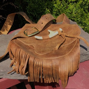 May include: A brown leather satchel bag with fringe detailing and a gold buckle. The bag has a strap and a bone-like handle.