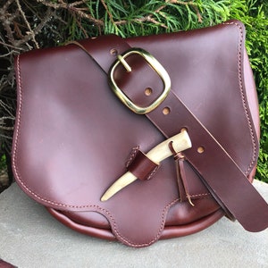 May include: A brown leather satchel bag with a brass buckle and a bone handle. The bag has a flap closure and a strap.
