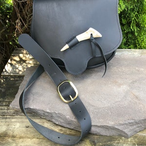 May include: A black leather pouch with a curved flap closure, featuring a light-colored antler toggle secured by a leather cord. The pouch is accompanied by a matching black leather belt that includes a polished brass buckle with a rounded frame.
