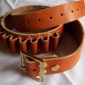 May include: A brown leather belt with a brass buckle and a cartridge loop. The belt is folded in half, showing the cartridge loop and the buckle.
