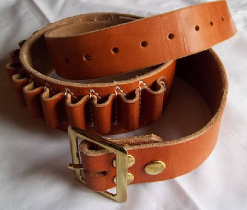 May include: A brown leather belt with a brass buckle and a cartridge loop. The belt is folded in half, showing the cartridge loop and the buckle.