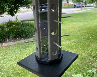 May include: A hexagonal bird feeder with a gray wood-grain finish, clear plastic panels, and a black base. It features multiple circular feeding ports and horizontal metal perches extending from the sides, topped with a black roof and hanging wire.