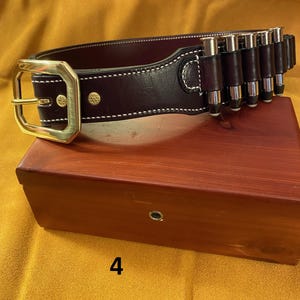 May include: A dark brown leather belt with a brass buckle and a cartridge loop. The belt is on a wooden box.