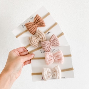 May include: Set of three nude-toned, textured, fabric hair bows on nude-colored elastic headbands. The bows are arranged in a stack, with the largest bow on top and the smallest bow on the bottom. The bows are all tied in a simple bow shape. The headbands are all made of a thin, elastic material. The text "Emilia Rose" is visible on the packaging.