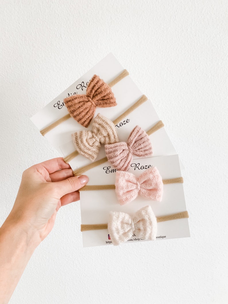 May include: Set of three nude-toned, textured, fabric hair bows on nude-colored elastic headbands. The bows are arranged in a stack, with the largest bow on top and the smallest bow on the bottom. The bows are all tied in a simple bow shape. The headbands are all made of a thin, elastic material. The text "Emilia Rose" is visible on the packaging.