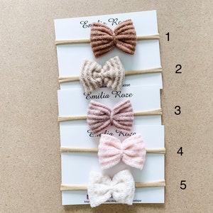 May include: Five different colored hair bows on a white card with the text "Emilia Roze" and numbers 1 through 5. The bows are made of a textured fabric and are arranged in a vertical line.
