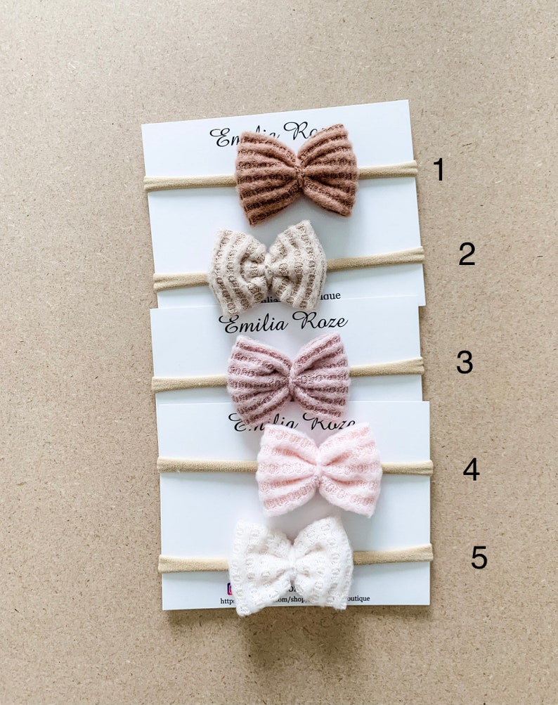 May include: Five different colored hair bows on a white card with the text "Emilia Roze" and numbers 1 through 5. The bows are made of a textured fabric and are arranged in a vertical line.