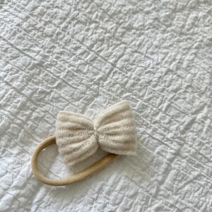 May include: A white knitted bow headband with a nude elastic band.