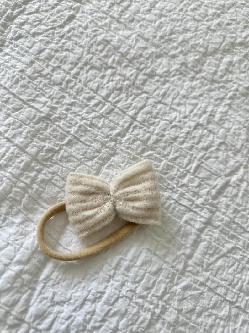May include: A white knitted bow headband with a nude elastic band.