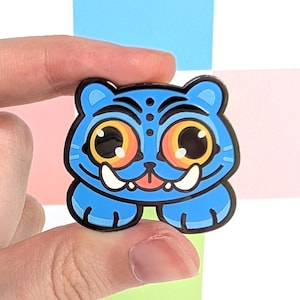 May include: A blue enamel pin depicting a cartoon tiger with large orange eyes, white tusks, and a black outline. The pin is held by a hand against a backdrop of pastel squares.