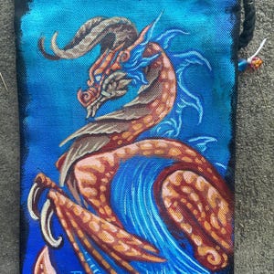 May include: A blue canvas bag with a hand-painted orange and blue dragon design. The dragon has a long, curled horn and a fierce expression. The bag has a drawstring closure and is signed "2024" in the bottom right corner.