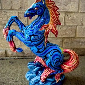 May include: A vibrant, hand-painted ceramic horse statue in a rearing pose. The horse features a blue body with intricate swirling patterns, a red and yellow mane and tail, and is mounted on a blue base. The statue is approximately 12 inches tall.
