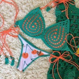 May include: A crocheted bikini top and bottom in a green and orange colour scheme. The top has a circular pattern and the bottom has a cherry print.