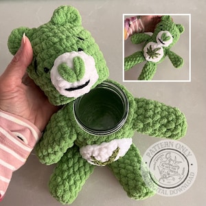 Pode incluir: Um urso verde de crochê com um focinho branco e um pote para o corpo. O urso tem um remendo branco no peito com um desenho de folha verde. A cabeça do urso é separada do corpo. O urso é feito à mão.