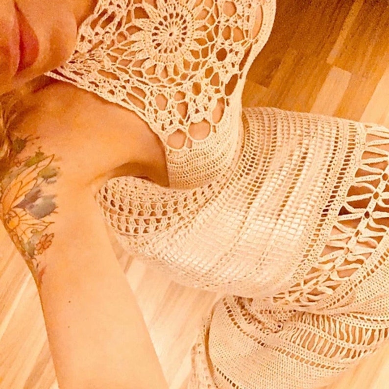 K&ouml;nnte beinhalten: Nahaufnahme eines geh&auml;kelten Kleides mit einem zarten Blumenmuster. Das Kleid ist aus hellbeigem Garn gefertigt und hat einen lockeren, flie&szlig;enden Sitz.