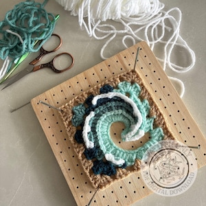 Pode incluir: Um motivo de crochê quadrado com um design de onda em espiral em tons de azul-petróleo, azul escuro e branco, preso a uma tábua de bloqueio de madeira com alfinetes de metal. Perto há fio azul-petróleo, uma agulha de crochê e tesouras vintage.