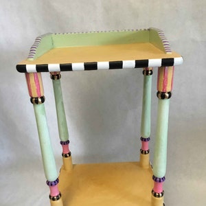 May include: A two-tiered side table with a green and yellow painted finish. The table has black and white checkerboard trim and pink, purple, and gold accents.