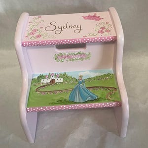 May include: A pink wooden step stool with a princess theme. The top is painted with the name "Sydney", a crown, and floral accents. The step features a painted scene of a princess in a blue gown near a castle.