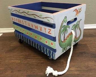 May include: A hand-painted wooden toy crate on wheels, featuring a blue interior and exterior accents. The crate is decorated with a green dinosaur, alphabet letters, and a rope handle. The crate is approximately 18 inches long, 12 inches wide, and 10 inches tall.