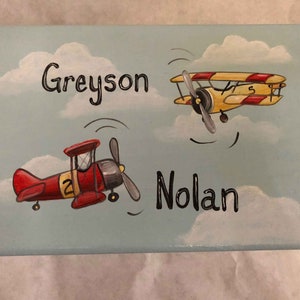 May include: A light blue wooden box with a painted sky scene. Two airplanes are painted on the box, one red and one yellow. The names Greyson and Nolan are written on the box.