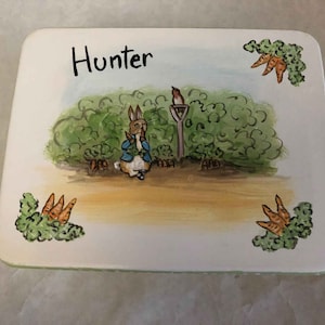 May include: A white ceramic tile with a green, brown, and orange illustration of Peter Rabbit from the children's book by Beatrix Potter. The tile is personalized with the name "Hunter" in black lettering.