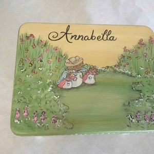 May include: A hand-painted wooden box with a yellow background and green grass. The box features a whimsical illustration of three mice in a field of flowers. The text "Annabella" is written in cursive on the box.