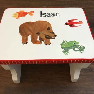 May include: White wooden step stool with a red and yellow border. The top of the stool is painted with a brown bear, a red bird, a green frog, and an orange fish. The name "Isaac" is written in black letters on the top of the stool.