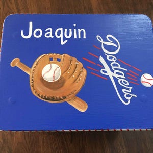 May include: A blue wooden step stool with a baseball glove, baseball, and bat painted on it. The stool is decorated with red and white stripes around the edge. The words "Joaquin Dodgers" are painted on the stool.
