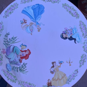 May include: A white round table top with hand-painted Disney princess characters. The table top features Cinderella, Ariel, Belle, and Jasmine. The characters are surrounded by floral designs.