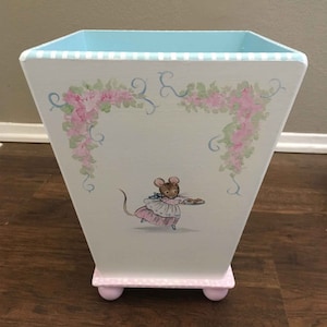 May include: A white painted wooden trash can with a light blue top and pink feet. The can is decorated with a hand-painted image of a mouse wearing a pink dress and carrying a plate of cookies. The top of the can has a white and light blue striped pattern.