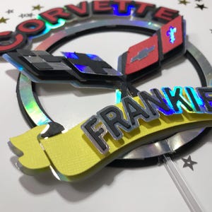 May include: A black and red Corvette logo with a checkered flag design and the word "FRANKIE" in a silver and blue iridescent font on a yellow banner. The logo is on a cake topper with a clear plastic stick.