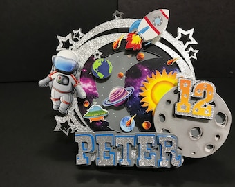 May include: A space-themed birthday cake topper with an astronaut, rocket, planets, and a sun. The design includes the name "PETER" and the number "12". The background features a galaxy design with silver glitter accents.