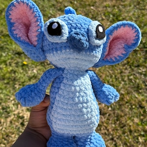 May include: A handmade blue crochet plush toy with large ears, pink inner ear details, and large black eyes. The plush has a textured body and is a character from a popular animated film.