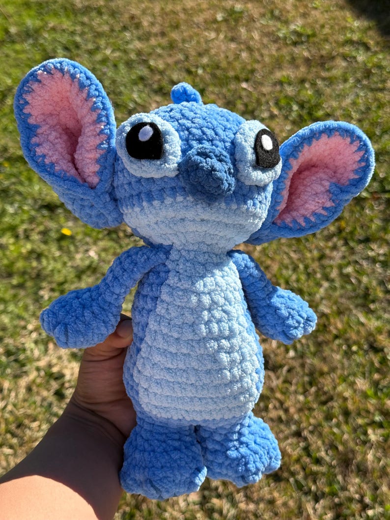 May include: A handmade blue crochet plush toy with large ears, pink inner ear details, and large black eyes. The plush has a textured body and is a character from a popular animated film.