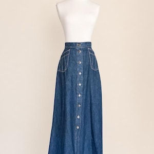May include: A high-waisted, floor-length denim skirt in a medium blue wash. The skirt features a vertical row of silver-toned metal buttons down the center front and two patch pockets on the hips, each accented with decorative vertical stitching.