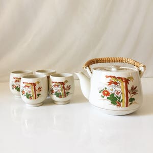 Può includere: Una teiera in porcellana bianca con un motivo a bambù e fiori e un manico intrecciato, insieme a quattro tazze da tè abbinate.