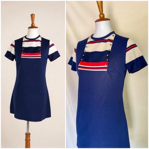 May include: A short-sleeved navy blue dress featuring a cream-colored chest panel with horizontal red and navy stripes. The front panel is accented with two vertical rows of small gold-toned buttons. The sleeves also feature matching stripes.