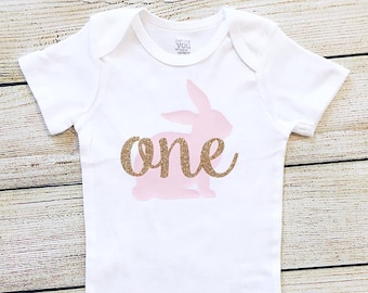 Puede incluir: Unisie blanco para bebé con una silueta de conejo rosa y texto de purpurina dorada que dice "one".