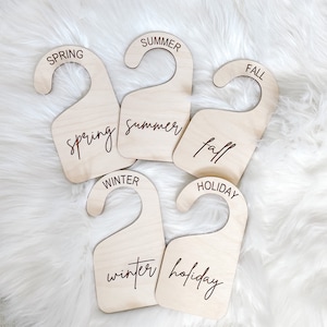 May include: Five wooden hangers with the words "Spring", "Summer", "Fall", "Winter", and "Holiday" written on them. The hangers are arranged in a semi-circle on a white fluffy surface.